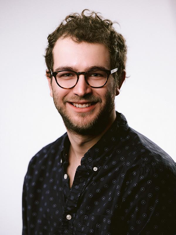 A smiling bearded man with curly dark hair wearing glasses and a dark patterned shirt