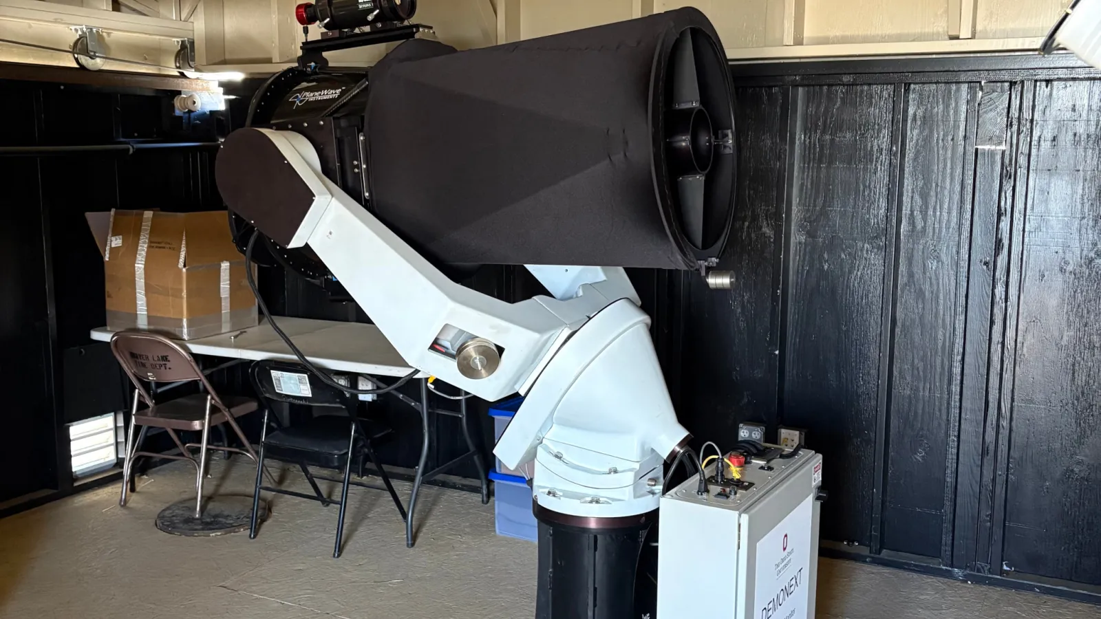 Black telescope on a white fork mount inside a roll-off roof enclosure.